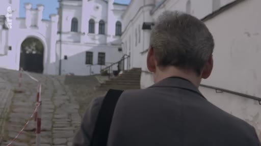 Un homme de dos se dirige vers une ancienne structure en pierre, possiblement un monastère ou un bâtiment historique, dans un cadre extérieur.