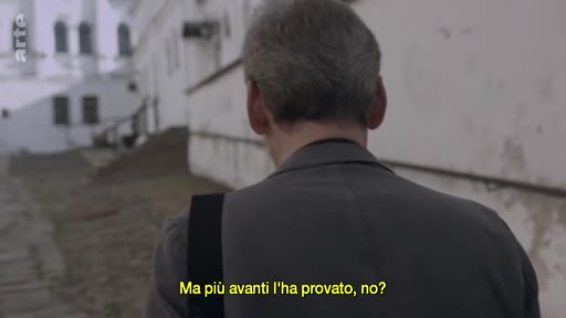 Un homme vu de dos marche le long d'un mur blanc dans une ruelle pavée, avec un sous-titre en italien affiché à l'écran.