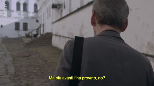 Un homme vu de dos marche le long d'un mur blanc dans une cour pavée, devant un bâtiment ancien avec des fenêtres cintrées. L'image est accompagnée d'un sous-titre en italien.