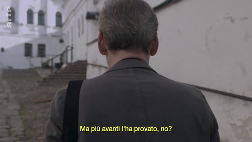 Un homme vu de dos marche le long d'un mur blanc dans une ruelle pavée, avec un bâtiment historique en arrière-plan. Une sous-titre en italien est visible à l'écran.