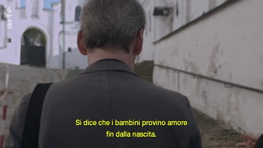 Un homme vu de dos marche dans une rue pavée devant un bâtiment blanc, accompagné d'un sous-titre en italien évoquant l'amour ressenti par les enfants dès la naissance.