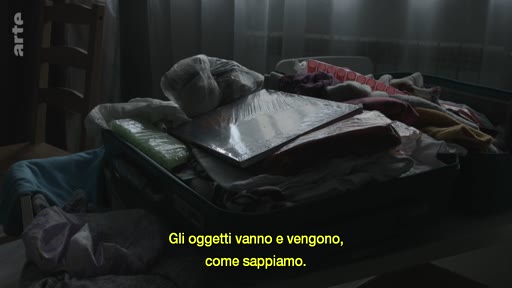 Une valise ouverte contenant divers objets personnels, des vêtements et des documents, illustrant le thème du déménagement ou du voyage. L'image est accompagnée d'un sous-titre en italien sur la nature éphémère des objets.