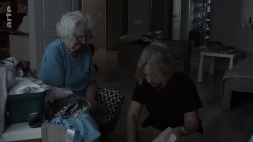 Une jeune femme blonde et une femme âgée aux cheveux blancs sont assises dans une pièce sombre, en train de trier des vêtements et des objets personnels, évoquant un moment de transmission ou de rangement familial.