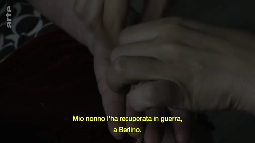 Un gros plan montre deux mains se tenant, illustrant un récit sur un objet récupéré par un grand-père à Berlin pendant la guerre. La scène souligne la transmission d'un héritage familial chargé d'histoire.