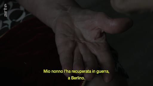Une main est filmée en gros plan, illustrant un témoignage sur un objet récupéré à Berlin pendant la guerre. Le sous-titre indique : 'Mio nonno l'ha recuperata in guerra, a Berlino'.