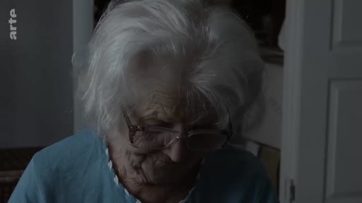 Gros plan sur une femme âgée aux cheveux blancs portant des lunettes, dans une séquence évoquant un trésor de famille sentimental.