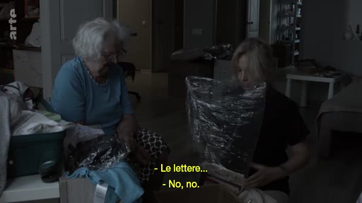 Une femme âgée aux cheveux blancs et une jeune femme blonde sont assises dans une pièce sombre en train de trier et d'emballer des objets dans des cartons, évoquant un déménagement ou un départ.