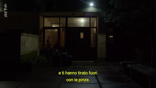 Une personne est assise devant l'entrée d'un bâtiment sombre la nuit, accompagnée de sous-titres en italien : 'e ti hanno tirato fuori con le pinze'.