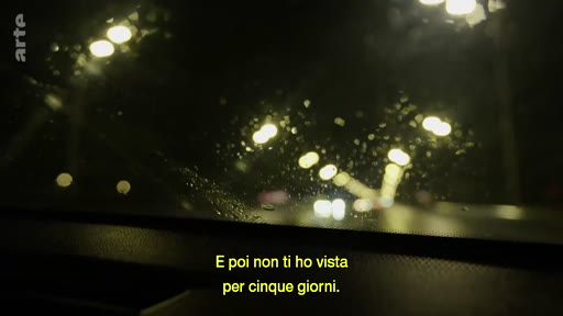 Vue subjective depuis le pare-brise d'une voiture circulant de nuit sous la pluie, avec des reflets de lumières urbaines et des sous-titres en italien.