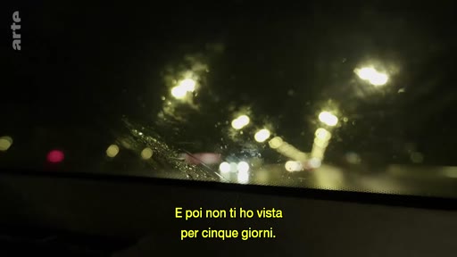 Vue subjective depuis l'intérieur d'une voiture circulant de nuit sur une route mouillée, avec des sous-titres en italien affichés à l'écran.