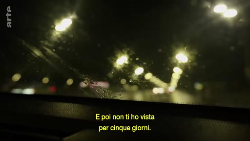 Vue subjective depuis le pare-brise d'une voiture circulant de nuit sur une route éclairée par des lampadaires, avec des sous-titres en italien.