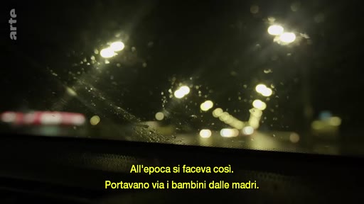Vue subjective depuis l'intérieur d'une voiture circulant de nuit sous la pluie, accompagnée d'un sous-titre évoquant la séparation des enfants et des mères. L'image illustre une atmosphère mélancolique ou dramatique liée à un témoignage sur des pratiques passées.