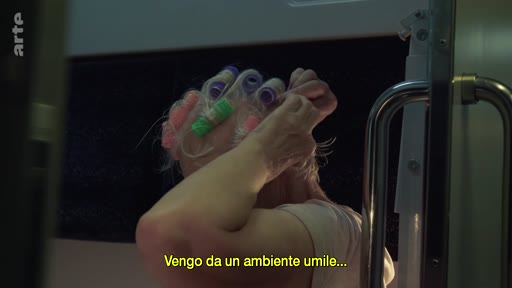 Une femme, vue de dos, ajuste ses bigoudis dans ses cheveux alors qu'elle voyage en train. Elle porte un haut clair et une sous-titre en italien est visible à l'écran.