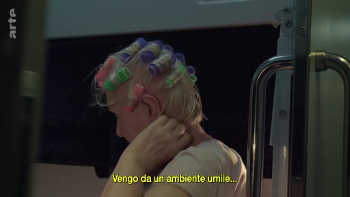 Une femme blonde portant des bigoudis colorés est assise dans un train, en train de se toucher les cheveux. Une incrustation de texte en italien indique qu'elle vient d'un milieu humble.