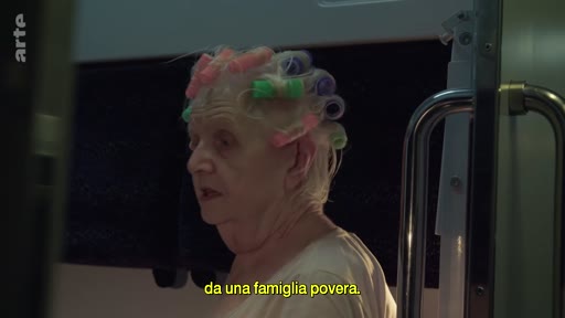 Une femme âgée portant des bigoudis dans les cheveux est filmée à l'intérieur d'un train, avec une légende en italien affichée à l'écran.