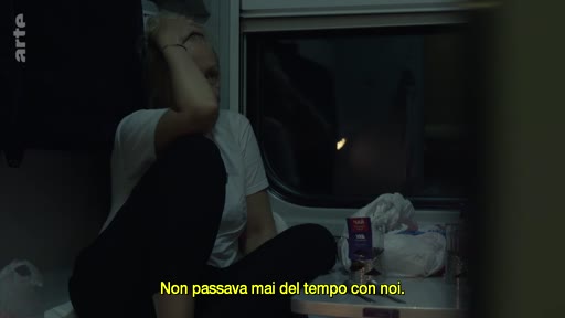 Une femme est assise dans un compartiment de train sombre, la main sur le front, dans une attitude pensive ou mélancolique. Des sous-titres en italien sont visibles à l'écran : 'Non passava mai del tempo con noi'.