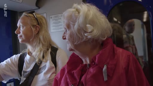 Une femme blonde et une femme âgée aux cheveux blancs observent le paysage depuis l'intérieur d'un train, lors d'un voyage en train.