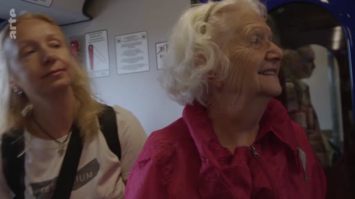 Deux femmes, dont une personne âgée aux cheveux blancs, sont assises côte à côte dans un wagon de train lors d'un voyage. Elles regardent par la fenêtre ou observent l'environnement intérieur du train.