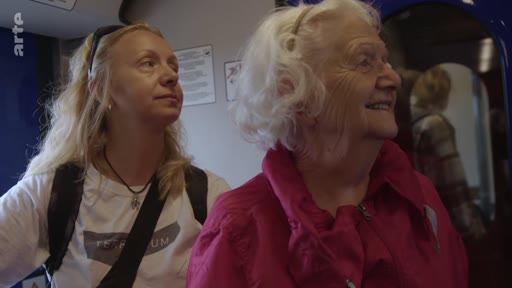 Une femme blonde et une femme âgée aux cheveux blancs voyagent ensemble à bord d'un train, regardant par la fenêtre. La scène illustre un moment de voyage ou de déplacement en transport en commun.