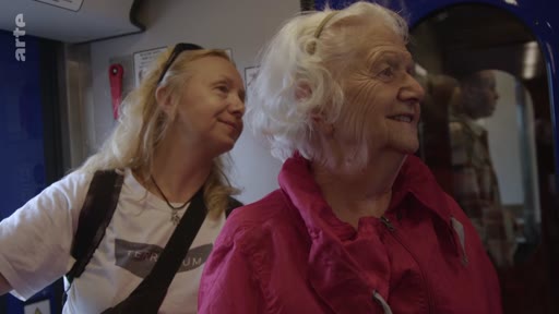 Une femme blonde portant un t-shirt blanc et un sac à dos accompagne une dame âgée aux cheveux blancs dans un wagon de train. La scène illustre un moment de voyage ou de déplacement, probablement dans le cadre d'un reportage sur l'assistance aux personnes âgées ou le transport ferroviaire.