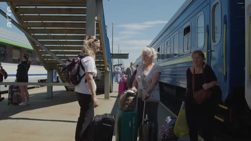 Des voyageurs, dont une femme âgée, attendent sur le quai d'une gare ferroviaire avec leurs valises à côté d'un train bleu. La scène montre le départ ou l'arrivée de passagers dans un contexte de voyage ou de déplacement de population.