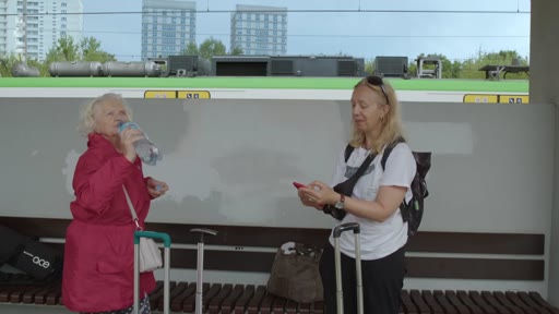 Une femme âgée en veste rouge et une femme plus jeune avec un sac à dos attendent sur le quai d'une gare, avec un train en arrière-plan. La femme âgée boit de l'eau tandis que la plus jeune consulte son téléphone portable.