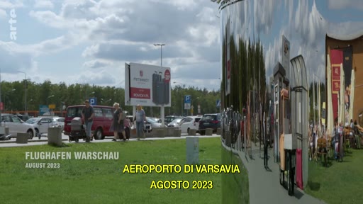Vue extérieure du parking et de l'entrée de l'aéroport de Varsovie en août 2023, avec des passagers marchant vers le terminal. Le texte à l'écran indique 'FLUGHAFEN WARSCHAU' et 'AEROPORTO DI VARSAVIA'.