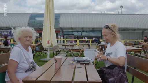 Une femme âgée aux cheveux blancs et une femme plus jeune sont assises à une table en bois en extérieur, devant un bâtiment de gare, lors d'une discussion. Elles semblent attendre ou se reposer dans un espace public aménagé avec des bancs et un parasol.