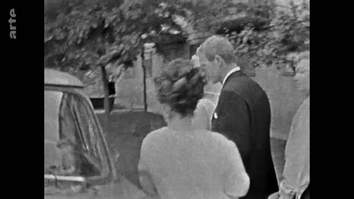 Le prince Philip, duc d'Édimbourg, est filmé de profil lors d'une apparition publique en extérieur, entouré de membres de la famille royale britannique. Cette image d'archive en noir et blanc montre le prince en costume sombre, marchant près d'une voiture.