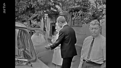 Une scène d'archive en noir et blanc montrant un homme en costume ouvrant la portière d'une voiture pour une femme, sous le regard d'un autre homme en chemise. Cette séquence illustre des moments de vie quotidienne ou de mariage dans un contexte historique.