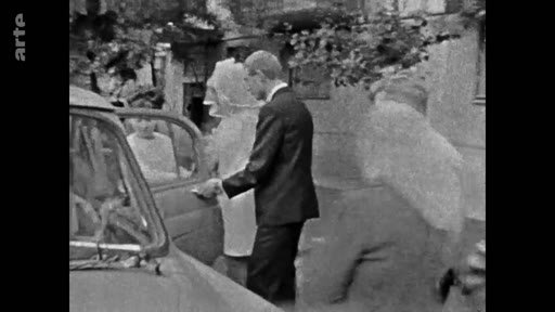 Une scène d'archives en noir et blanc montrant un homme en costume ouvrant la portière d'une voiture pour une femme portant une robe de mariée et un voile, probablement lors d'un mariage.