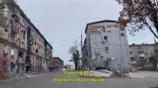 Vue d'une rue bordée d'immeubles résidentiels gravement endommagés par les combats en Ukraine, illustrant les destructions urbaines liées au conflit. Le sous-titre évoque le lieu de naissance et de croissance de la personne qui témoigne.