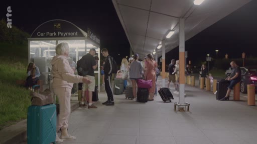 Des voyageurs avec leurs valises attendent près d'une borne de paiement de parking, identifiée par le texte 'Car Park Payment', lors d'une arrivée nocturne.