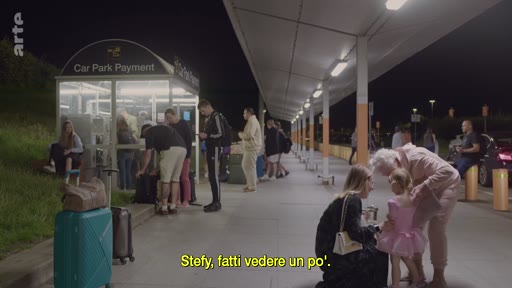 Une grand-mère retrouve sa petite-fille vêtue d'un tutu rose devant une borne de paiement de parking à l'extérieur, lors d'une arrivée à Londres. La scène montre des voyageurs avec leurs bagages dans un environnement nocturne.
