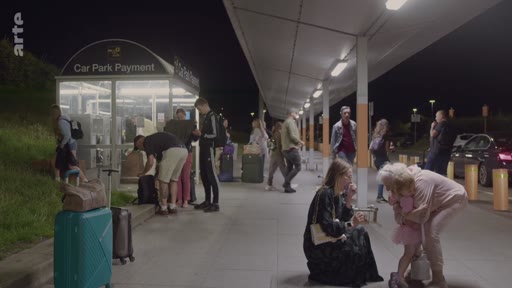 Des voyageurs avec leurs bagages attendent près d'une borne de paiement de parking, marquée 'Car Park Payment', dans une zone de dépose-minute la nuit.
