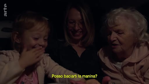 Une petite fille, sa mère et sa grand-mère sont assises ensemble dans une voiture, partageant un moment affectueux. La petite fille demande à embrasser la main de sa grand-mère, illustrant une scène de complicité intergénérationnelle.