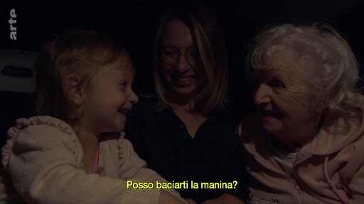 Une petite fille, sa mère et sa grand-mère sont assises ensemble dans une voiture, échangeant des gestes affectueux. La scène capture un moment d'intimité familiale lors d'un trajet nocturne, illustré par la question sous-titrée 'Posso baciarti la manina?'.