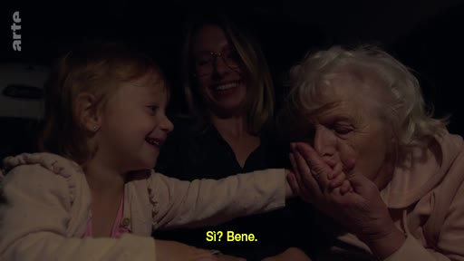 Trois générations de femmes, une petite fille, sa mère et sa grand-mère, sont réunies dans une voiture lors d'un moment de complicité familiale. La grand-mère embrasse la main de l'enfant tandis que la mère sourit, illustrant une scène de vie quotidienne et de transmission intergénérationnelle.