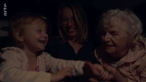 Trois générations de femmes, une petite fille, sa mère et sa grand-mère, sont réunies dans une voiture lors d'un trajet nocturne. Elles interagissent avec tendresse, illustrant un moment de complicité familiale.