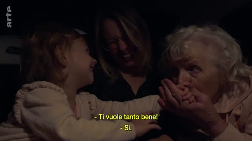 Une petite fille, sa mère et sa grand-mère sont assises ensemble dans une voiture, partageant un moment d'affection et de complicité. La grand-mère embrasse les mains de l'enfant tandis que la mère sourit, illustrant une scène de vie familiale intime.