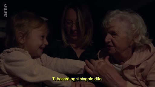 Une petite fille, sa mère et sa grand-mère sont assises ensemble dans une voiture, partageant un moment d'affection. La scène illustre une interaction familiale intime, accompagnée d'un sous-titre en italien.