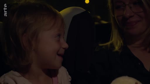 Une jeune enfant sourit en regardant sa mère dans l'habitacle d'une voiture, lors d'un trajet nocturne. La scène illustre un moment de complicité familiale.