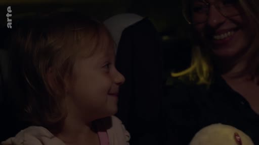 Une petite fille et sa mère partagent un moment de complicité et de tendresse lors d'un trajet en voiture, filmées dans une ambiance intime et sombre.