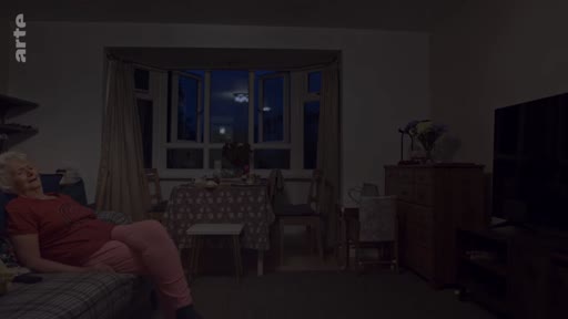 Une femme âgée est assise sur un canapé dans un salon sombre, avec une vue sur une fenêtre en soirée. L'ambiance est calme et domestique, typique d'un reportage sur la vie quotidienne des seniors.
