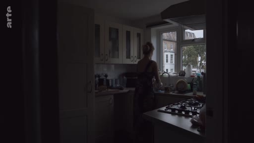 Une femme se tient dans une cuisine à la lumière tamisée, effectuant des tâches ménagères près de la fenêtre. L'ambiance est calme et domestique, typique d'un intérieur résidentiel.