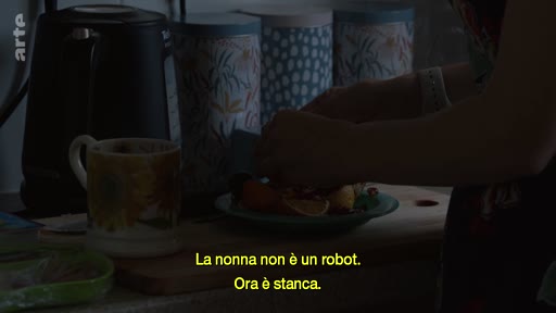 Une personne prépare des fruits sur une planche à découper dans une cuisine, accompagnée de sous-titres en italien évoquant la fatigue d'une grand-mère. La scène illustre un moment de vie quotidienne et domestique.