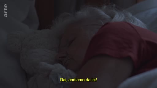 Une personne âgée aux cheveux blancs est allongée dans un lit, en train de dormir, avec un sous-titre en italien affiché à l'écran.