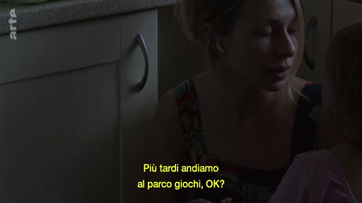 Une mère blonde s'adresse à son enfant dans une cuisine sombre, avec des sous-titres en italien à l'écran. La scène évoque un moment de complicité familiale et de vie quotidienne.