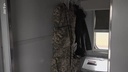 Vue intérieure d'un compartiment de train lors d'un voyage vers Marioupol, montrant des vêtements militaires suspendus et des objets personnels sur une tablette. Ce plan illustre le quotidien des déplacements dans la zone de conflit en Ukraine.