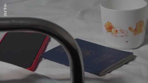 Un passeport ukrainien bleu est posé sur un lit, accompagné d'un téléphone portable et d'un gobelet illustré, dans le cadre du documentaire Journal d'un voyage à Marioupol.